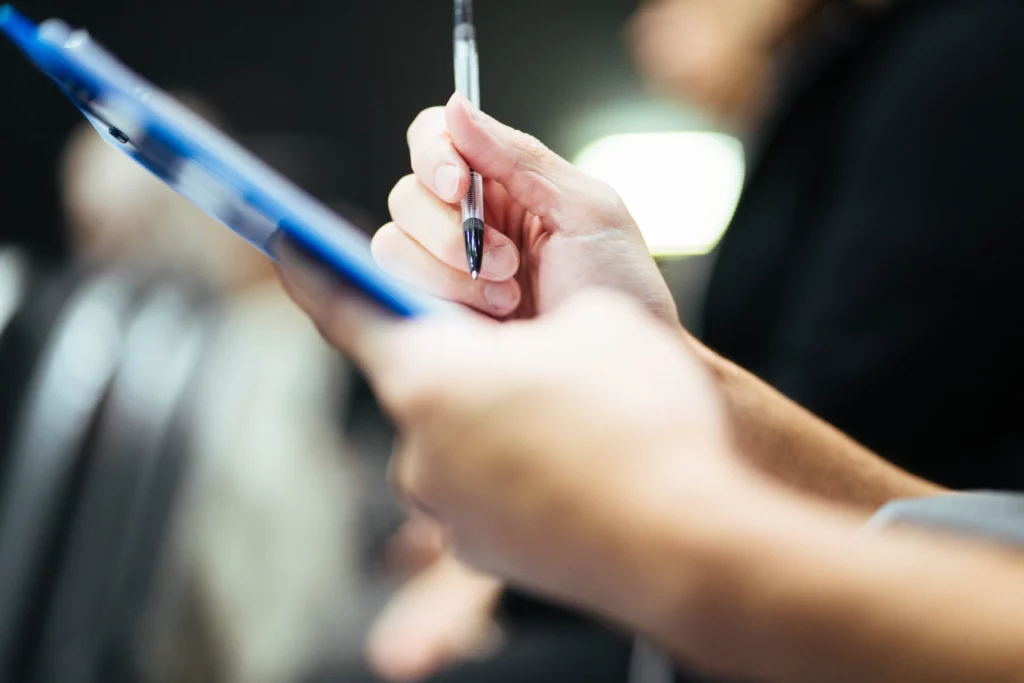 Hand mit Stift notiert Prüfergebnisse auf einer Klemmmappe