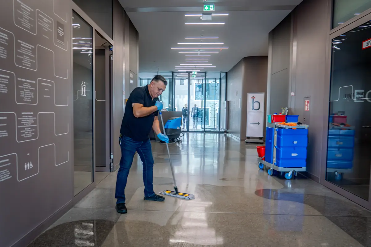 Unterhaltsreinigung: Reinigungskraft von ML Facility wischt den Flur eines modernen Bürogebäudes